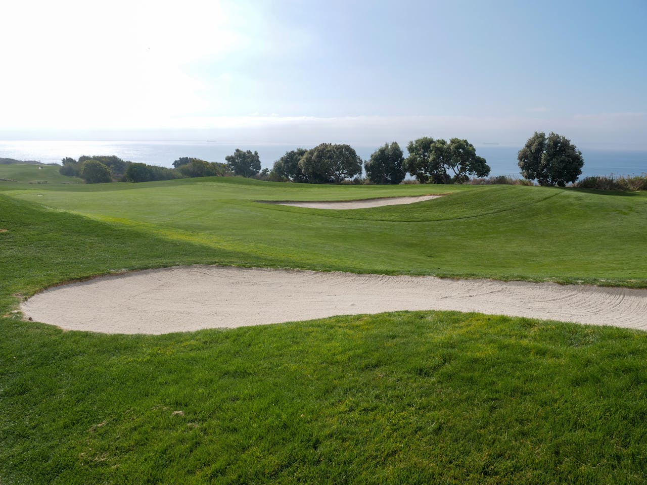Beautiful golf course with sand trap and ocean view under a clear sky.